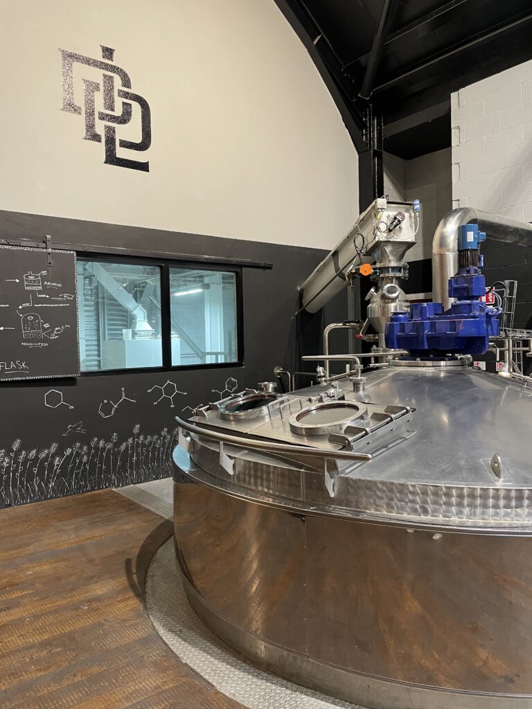 Industrial distillery interior with a large stainless steel vat in the foreground. Chalkboard wall with chemistry diagrams and a window behind. Modern and sleek ambiance.