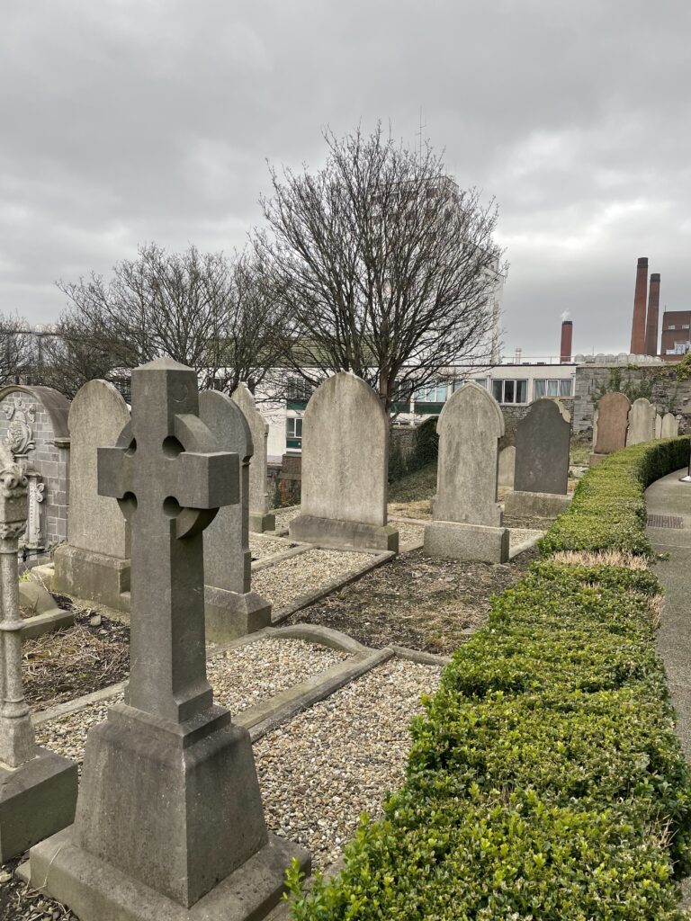 Cemetery landscape with gravestones and Celtic cross. Overcast sky above old tombstones, greenery, and bare trees.
