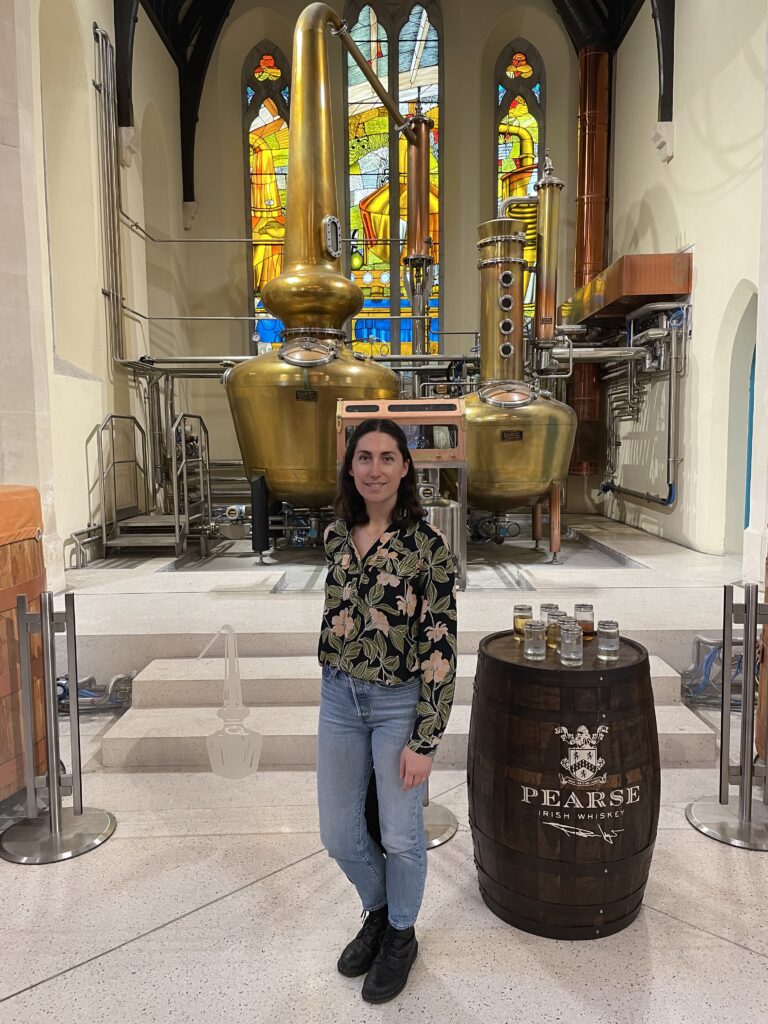 A woman in a floral blouse stands inside a distillery with tall brass distillation equipment and colorful stained glass windows. A barrel labeled "Pearse" holds small tasting glasses.