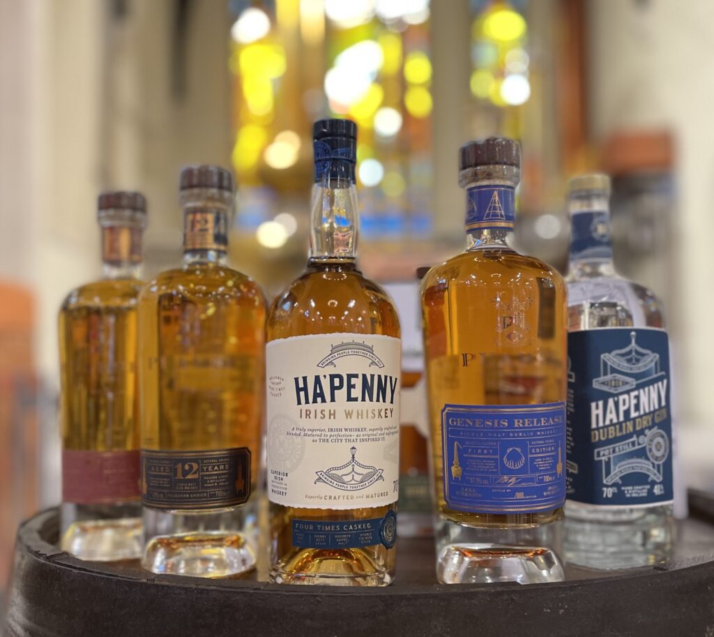 Close-up of a selection of Ha’Penny Irish spirits displayed on a wooden barrel, including bottles of Irish whiskey and Dublin dry gin. The bottles are sharply focused against a softly blurred background with warm, colorful lighting, showcasing premium offerings from Dublin distilleries.