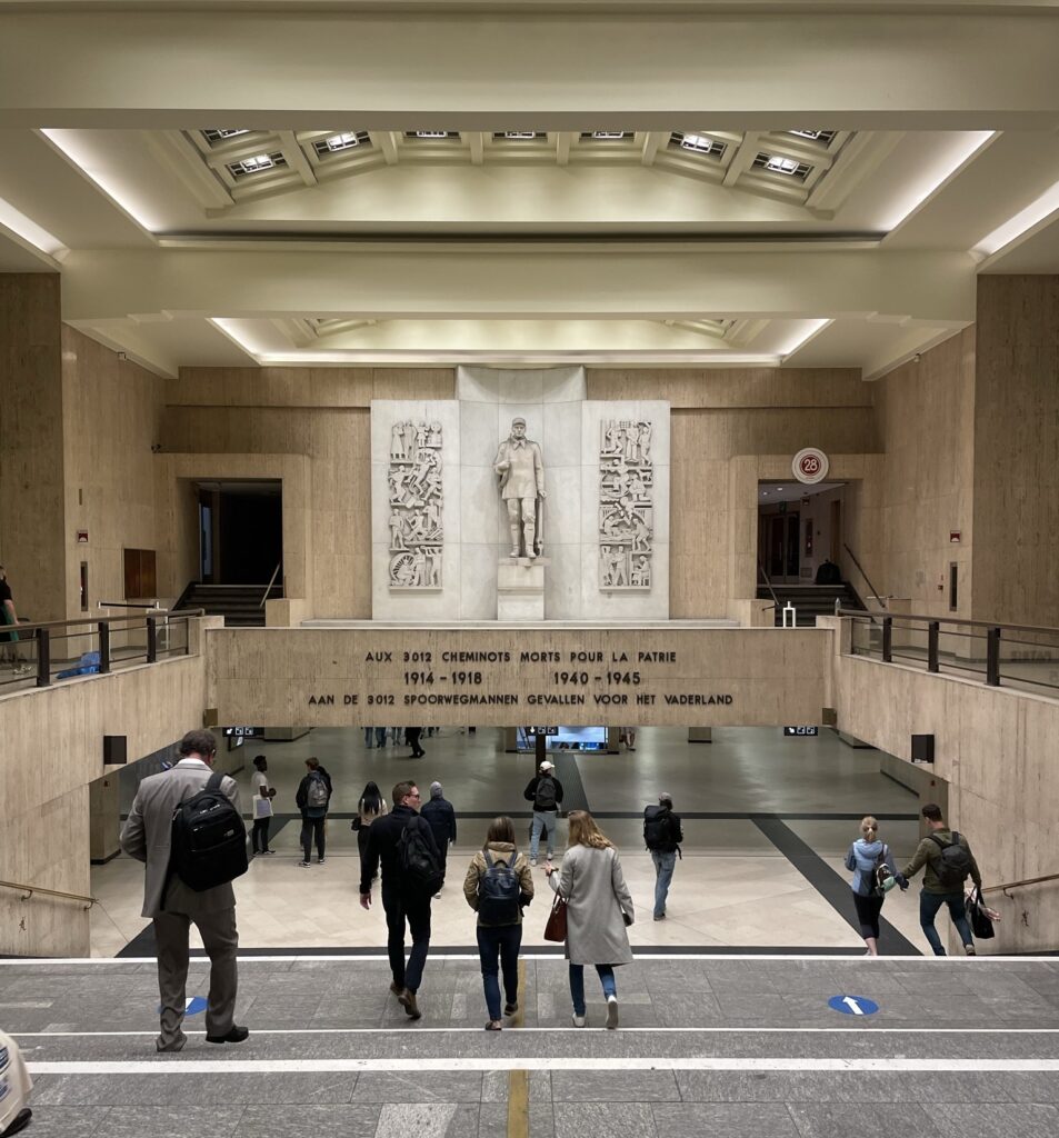 The grand hallway of Brussels Central train station with a large memorial featuring a soldier statue. People walk below, as they rush to and from their trains in Brussels' main transit hub.
