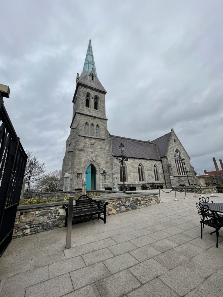 A stone church with a tall spire under a cloudy sky. The building has arched windows, a teal door, and is surrounded by a paved area with benches.