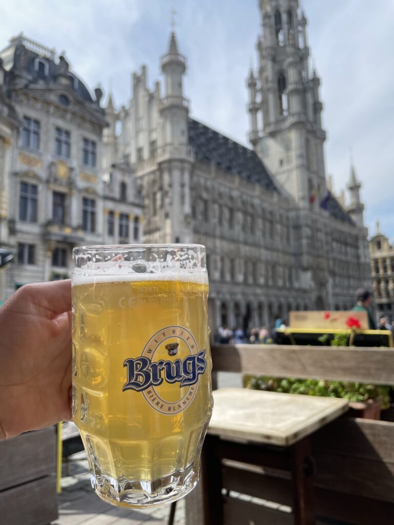 A hand holds up a glass of Brugs Witbier, a pale Belgian wheat beer, against the backdrop of ornate historic buildings in Brussels' Grand Place. The sunlight highlights the beer’s golden color as people enjoy the outdoor café setting.
