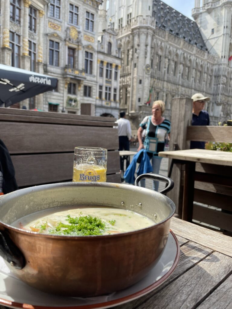 A steaming bowl of creamy soup garnished with green herbs in a metal pot sits on a wooden table. A glass of beer is beside it. Historic buildings and people walking provide a lively European street scene in the background.