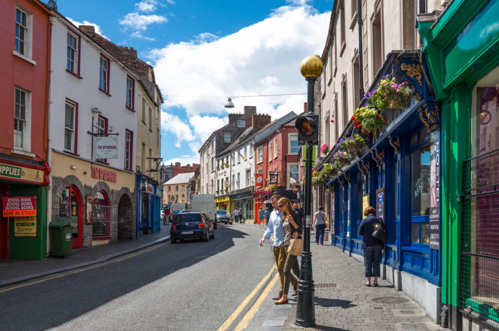 A lively street in Kilkenny, Ireland features rows of colorful shopfronts with hanging flower baskets and bright painted facades. Pedestrians walk along the narrow sidewalk while cars move down the curved road under a sunny sky with scattered clouds. The scene captures the charm of the historic town center with its mix of small businesses and vibrant street life.