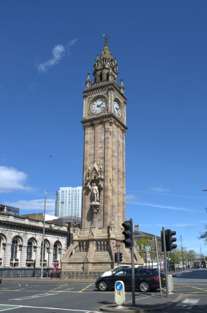 The tall Prince Albert Memorial clock tower stands against a clear blue sky in an urban setting. Cars and traffic lights are visible at its base, conveying a busy city atmosphere.