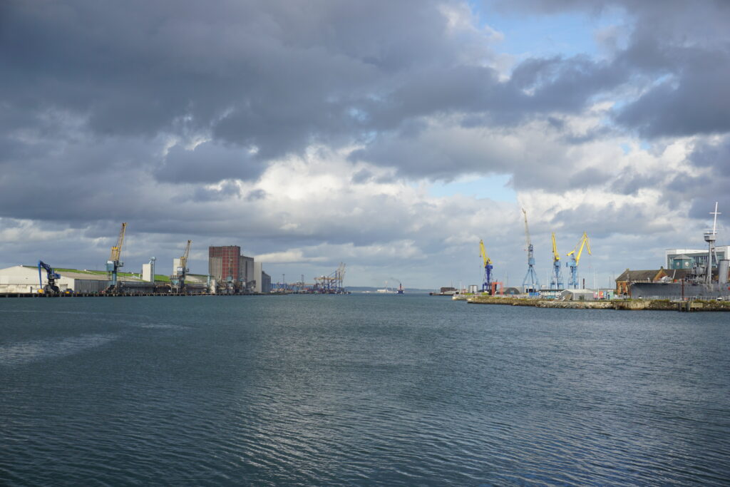 A wide view of Belfast Harbour shows calm water bordered by industrial docks lined with warehouses, cranes, and shipping equipment. Yellow and blue cranes rise against a backdrop of heavy gray clouds with a few patches of blue sky. The scene captures the scale of the working port and its mix of modern and older waterfront structures.