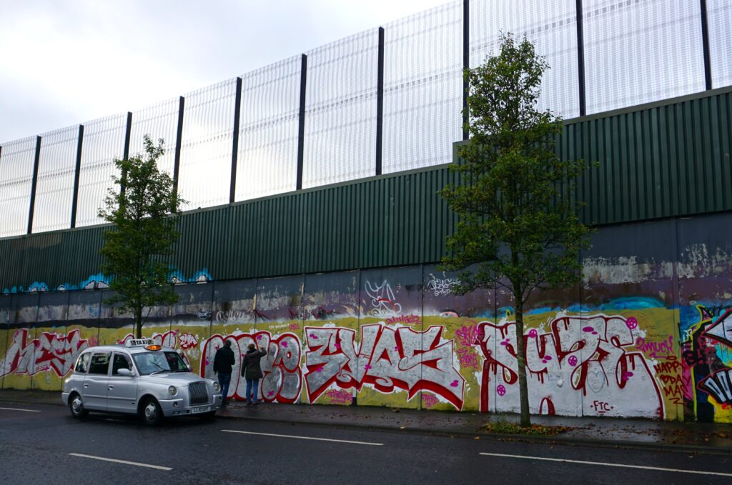 Urban street scene with a tall fence and colorful graffiti. A silver taxi is parked by the curb with two people standing nearby. Overcast sky, two trees.