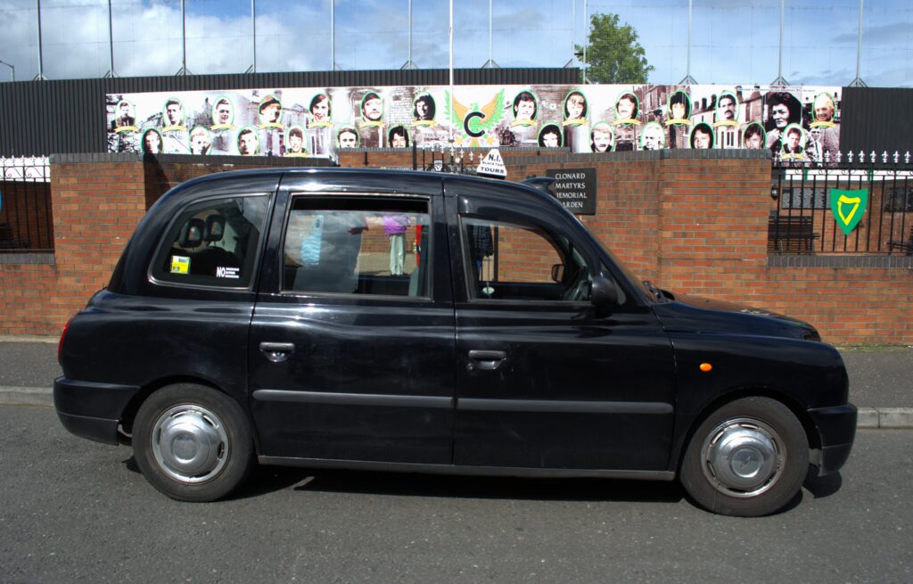 Black taxi cab parked in front of a brick wall adorned with a colorful mural featuring various faces. The sky is partly cloudy.