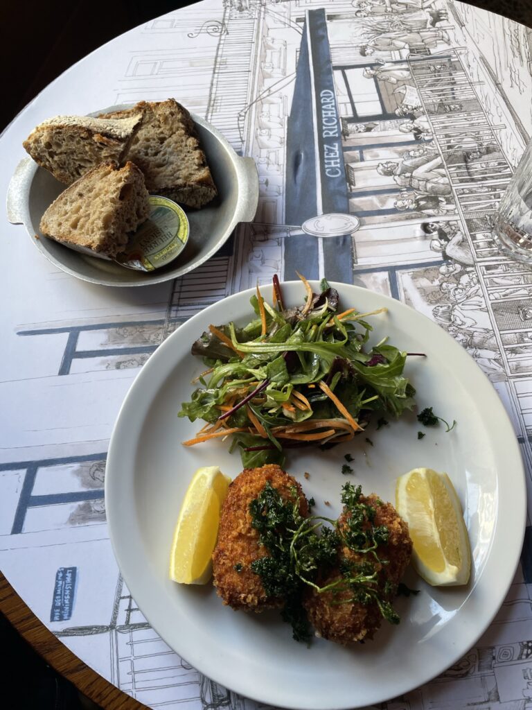 A white plate holds two golden croquettes topped with fried herbs, served with lemon wedges and a small green salad of mixed leaves and shredded carrots. Beside it, a small dish of rustic brown bread slices and butter sits on a placemat illustrated with a café scene labeled “Chez Richard.”