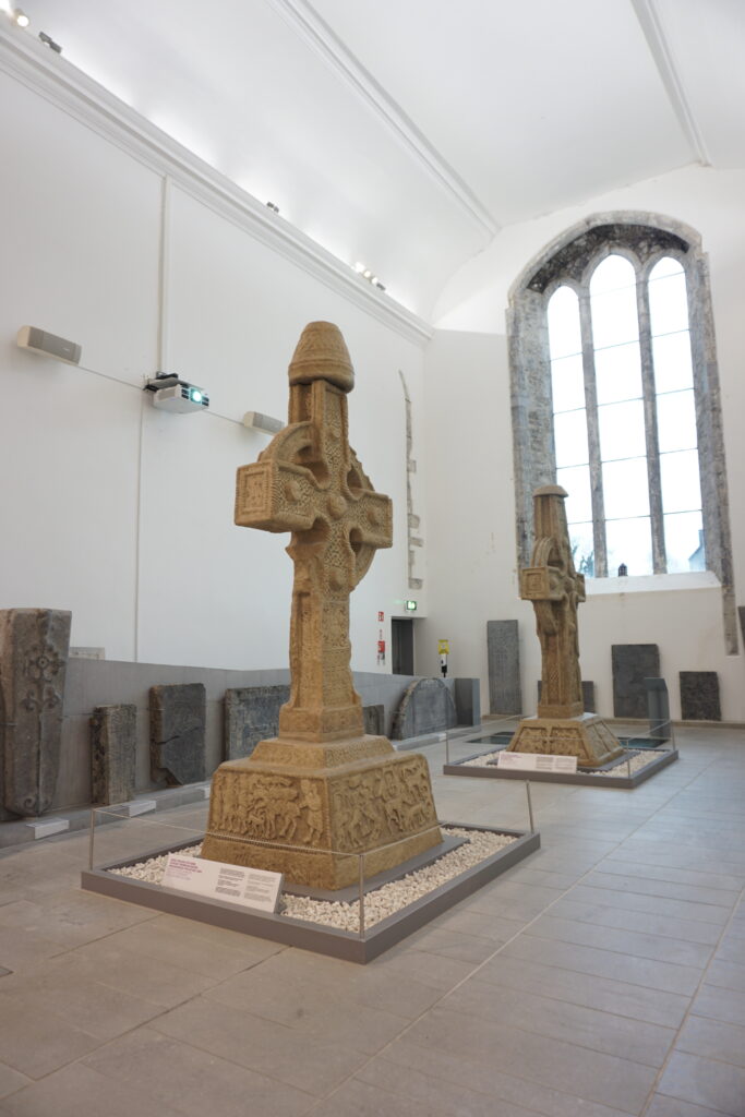 Two intricately carved stone Celtic crosses are displayed in a bright museum room with tall arched windows, evoking a sense of historical reverence.