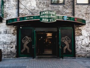 Entrance to the historic Jameson Distillery Bow St. in Dublin, featuring weathered brick walls, green signage, and wooden whiskey barrels outside. A green awning marks the doorway to one of the most famous Dublin distilleries.