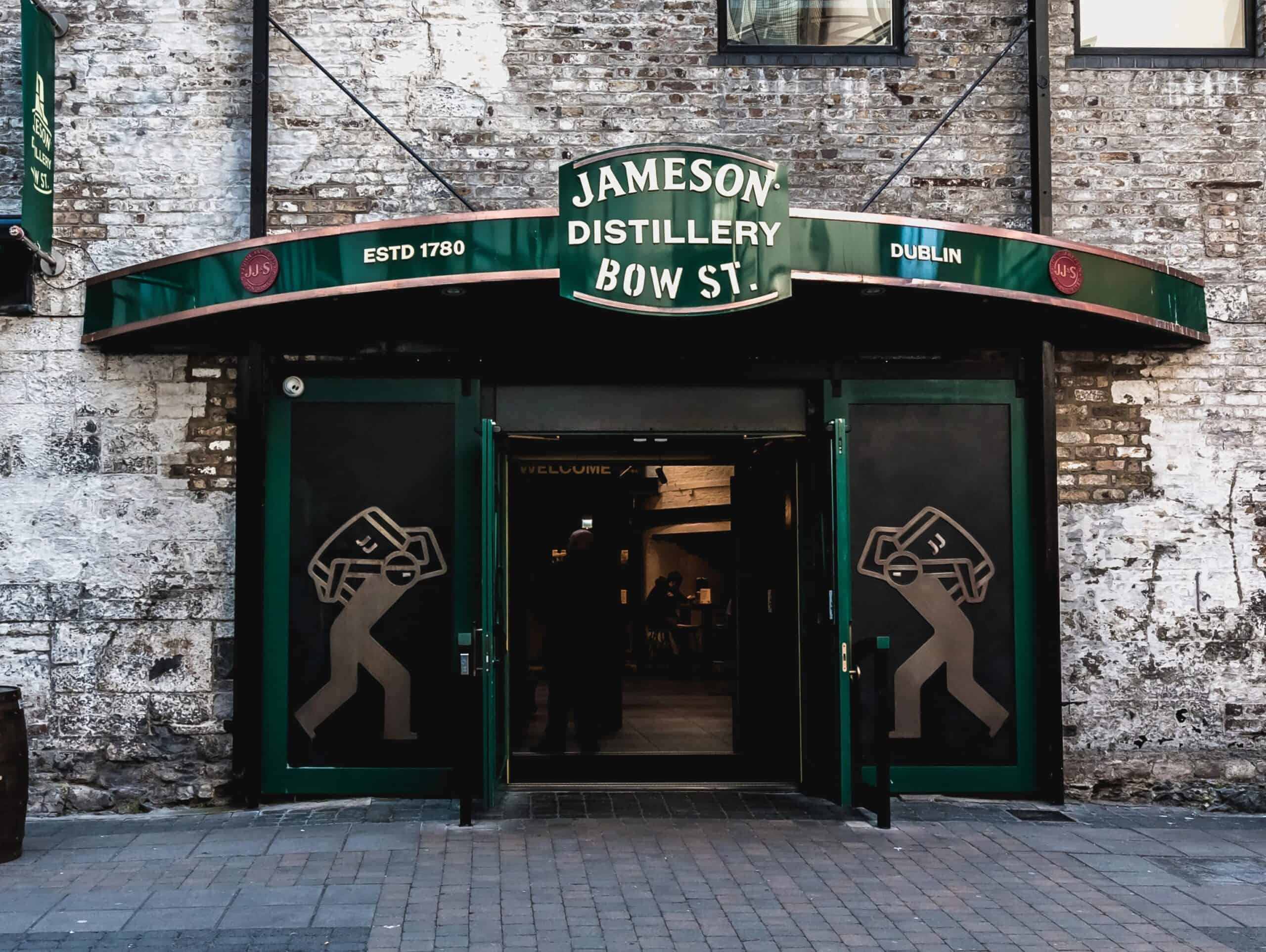 Entrance to the historic Jameson Distillery Bow St. in Dublin, featuring weathered brick walls, green signage, and wooden whiskey barrels outside. A green awning marks the doorway to one of the most famous Dublin distilleries.