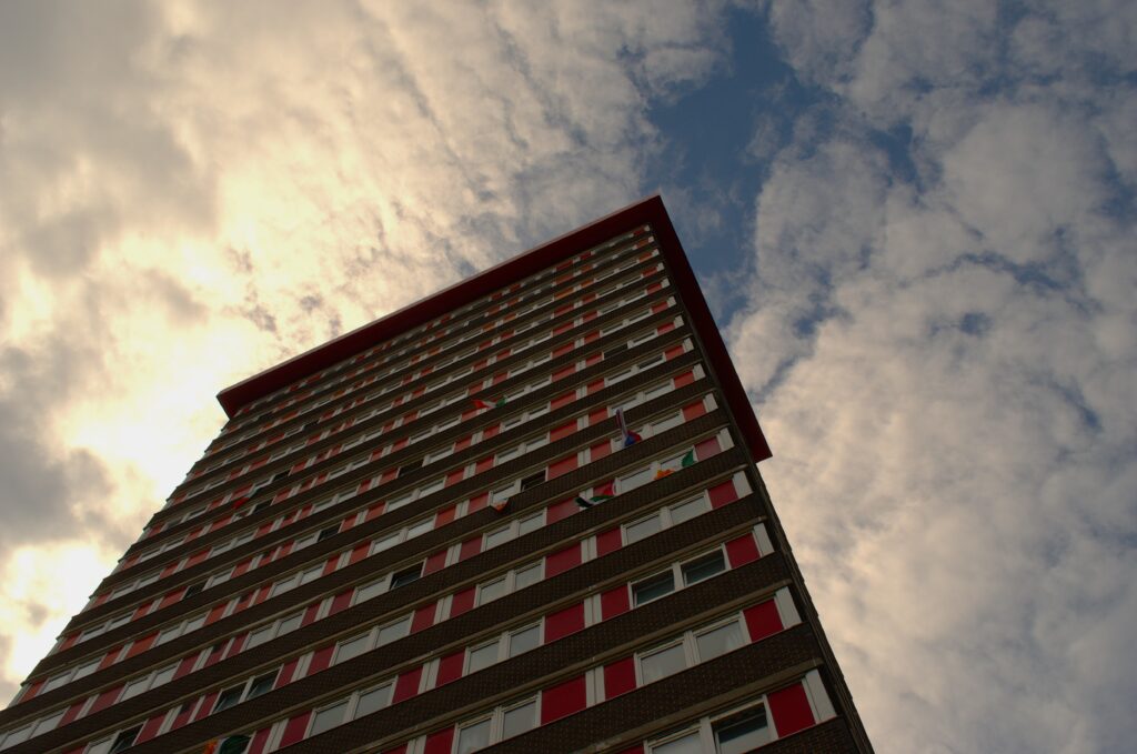 Tall building with red and white facade viewed from below. Above, the sky is partly cloudy, creating a dramatic and towering perspective.