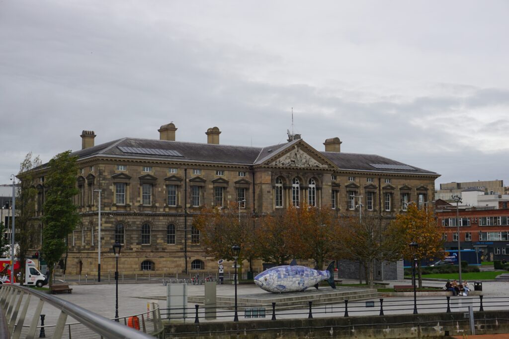 Historic stone building with arched windows, surrounded by trees with autumn leaves. A mosaic fish sculpture sits in the foreground. Overcast sky.