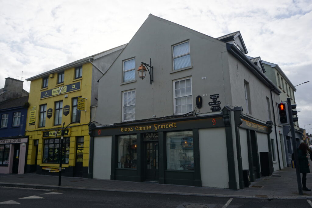 A street corner in Kilkenny, Ireland, featuring a light gray building with the shop “Siopa Cúinne Sincell” on the ground floor and shuttered windows above. Next to it stands a bright yellow storefront called “the zip yard,” adding contrast to the row of colorful buildings. A pedestrian waits near a red crossing signal under an overcast sky.