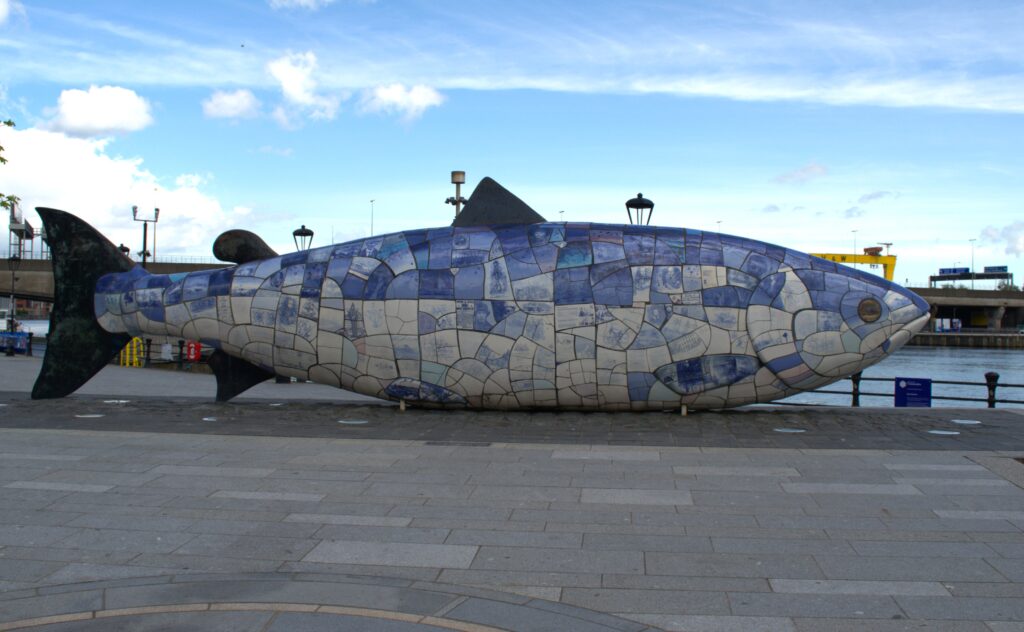 Colorful ceramic mosaic sculpture of a large fish on a paved walkway by the water. Blue sky and distant buildings create a calm urban setting.