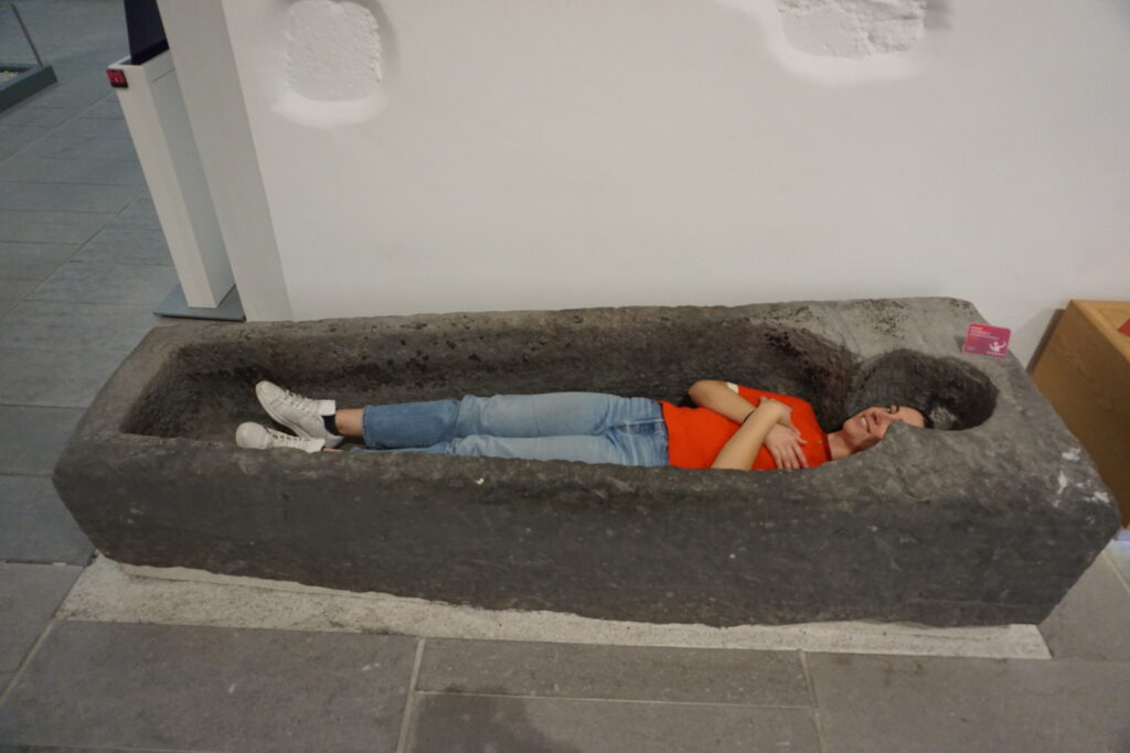 The author dressed in jeans and a read short sleeved shirt lies comfortably in an ancient stone sarcophagus at the Kilkenny Medieval Mile Museum, conveying a playful and relaxed mood in a museum setting.