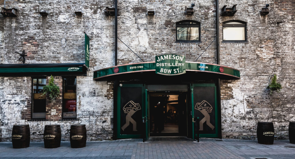 Entrance to the historic Jameson Distillery Bow St. in Dublin, featuring weathered brick walls, green signage, and wooden whiskey barrels outside. A green awning marks the doorway to one of the most famous Dublin distilleries.