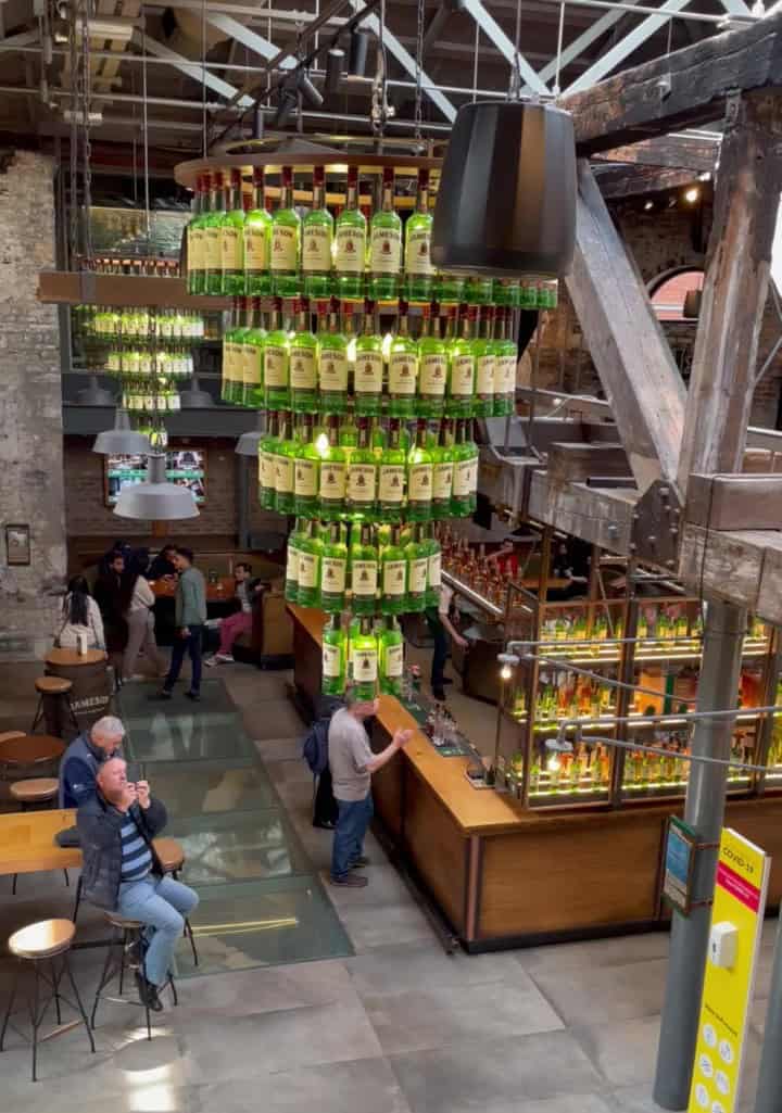 Interior of the Jameson Distillery in Dublin, showcasing a striking chandelier made from stacked Jameson whiskey bottles hanging above a bar. Visitors enjoy drinks and take photos in the lively space, highlighting the atmosphere typical of popular Dublin distilleries.