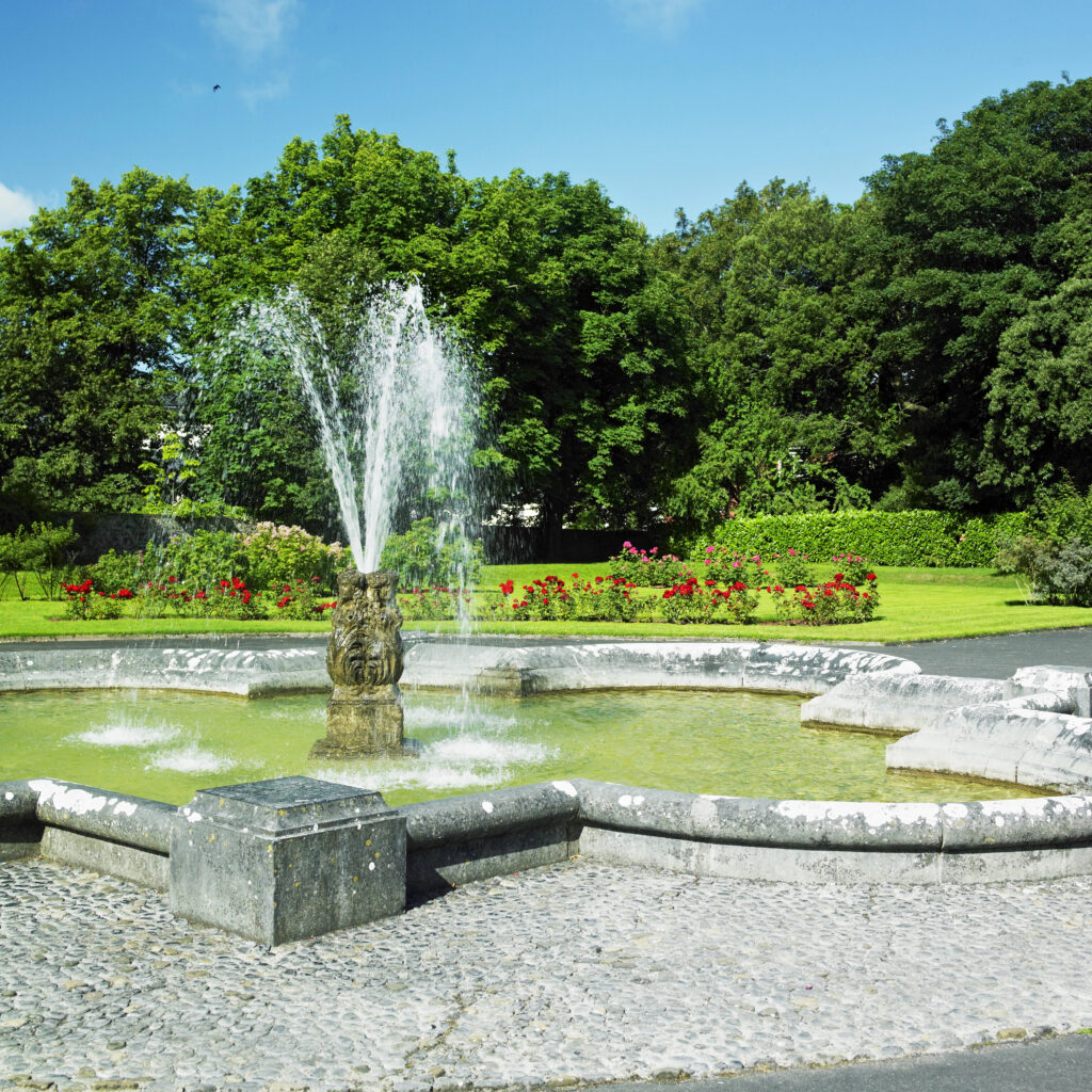 A fountain in the Kilkenny Castle gardens with water spraying upwards is set amid vibrant gardens, with red flowers and lush greenery under a clear blue sky, conveying a serene summer atmosphere.