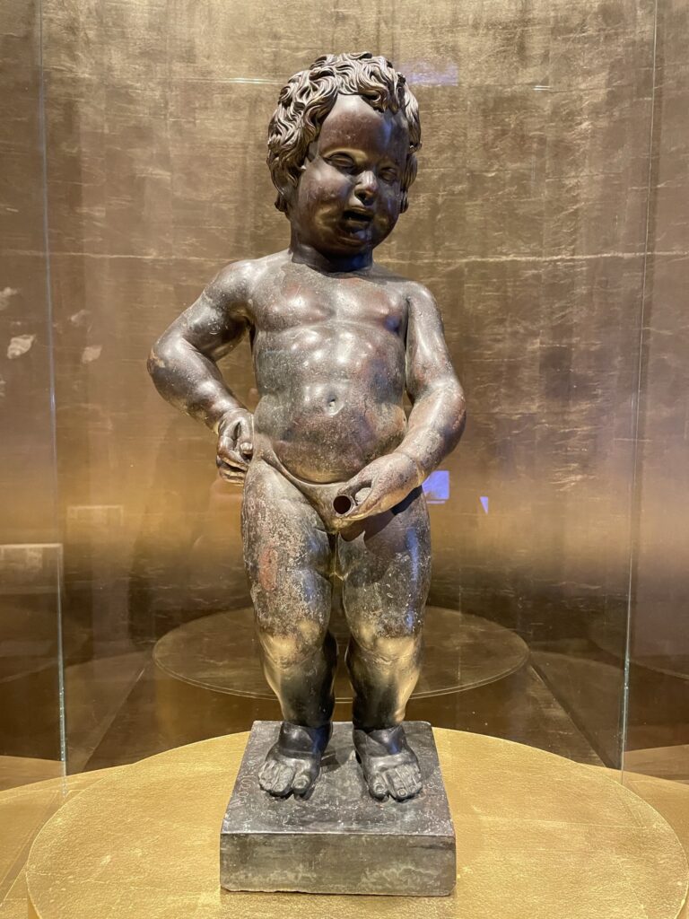 Bronze statue of a child with curly hair, standing with one hand on hip and other at stomach. The background is a textured golden surface.