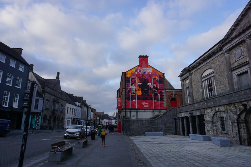A vibrant mural on a building's side contrasts with gray stone architecture on a quiet street. A lone person walks, creating a calm, urban atmosphere.