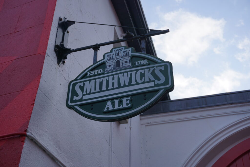 Sign for "Smithwick's Ale" with a castle emblem, hanging from a red and white building. The sky is partly cloudy, creating a classic pub atmosphere.