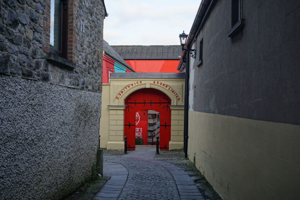 Narrow cobblestone alley between stone buildings leads to a red arched doorway marked "Smithwick & Sons Limited," conveying historic charm.