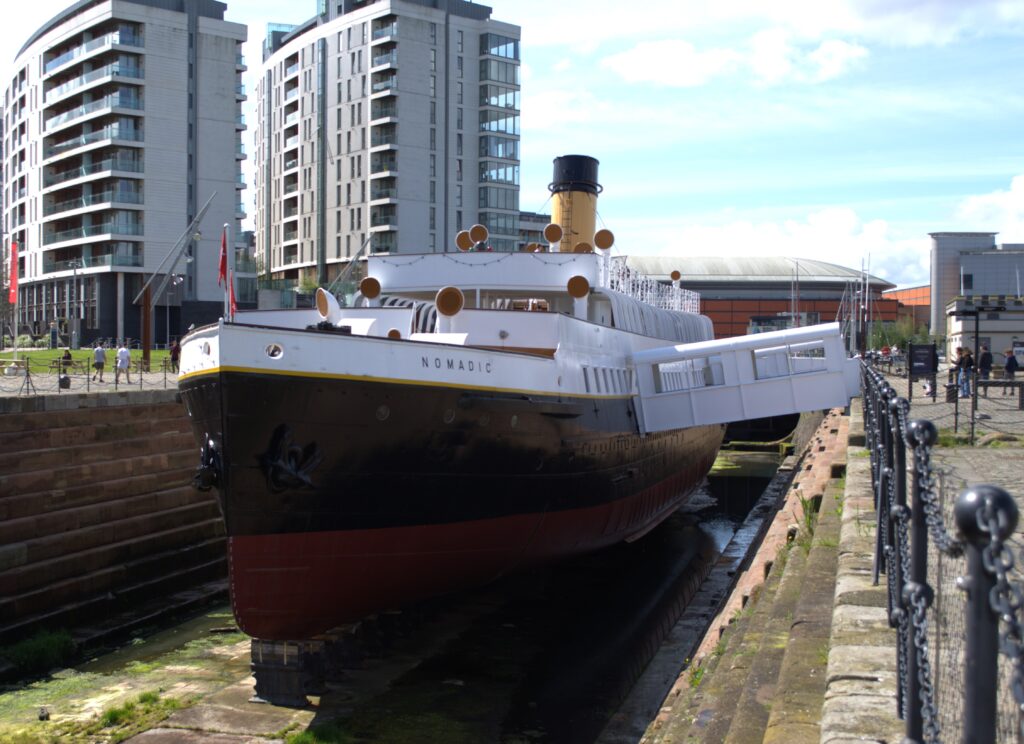 Historic ship Nomadic in dry dock, with modern buildings in the background. The ship's black hull and white upper structure shine under a sunny sky.