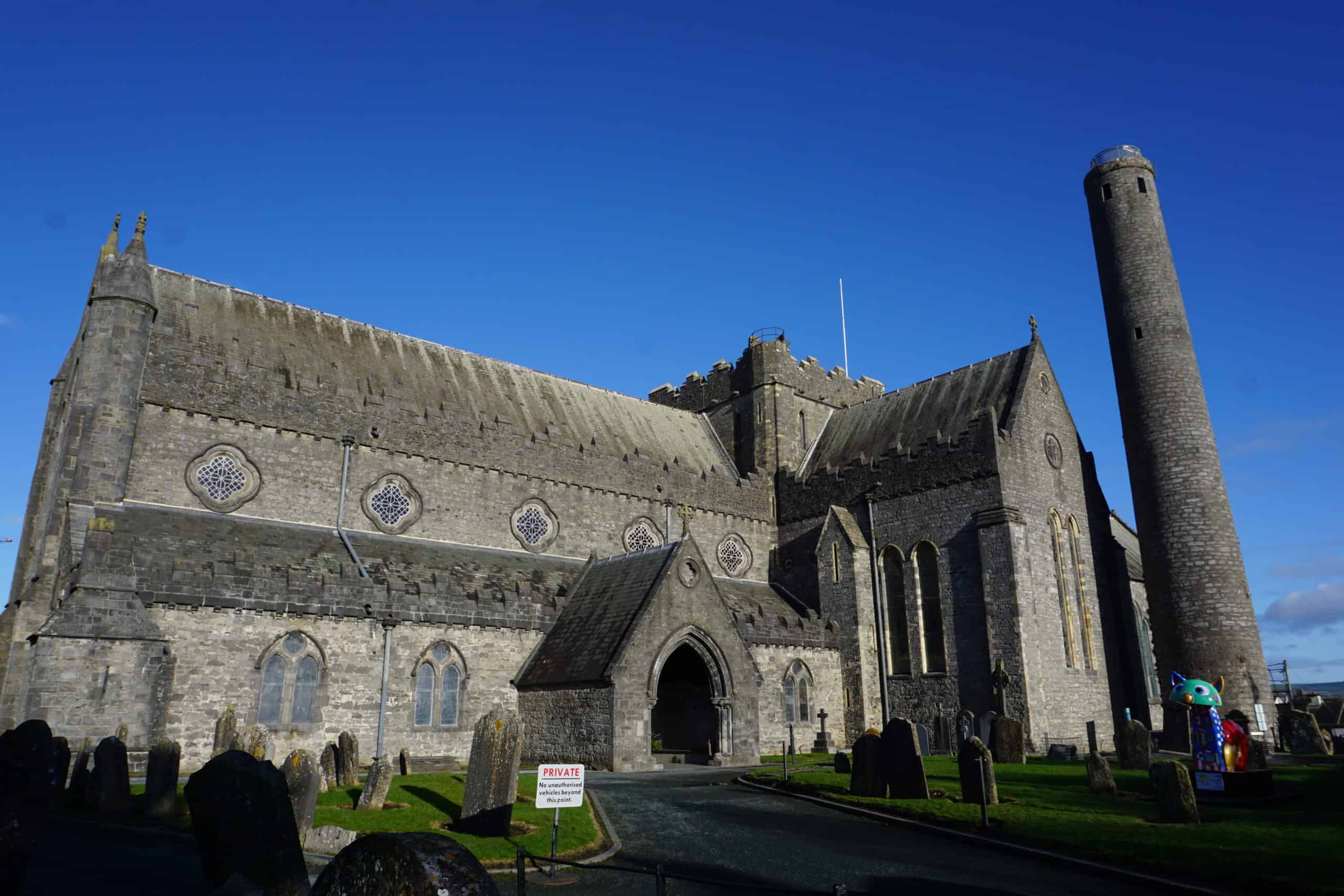 The large stone medieval church of Kilkenny City's St Canice's Cathedral stands under a bright blue sky, with a tall round tower rising beside it. Weathered gravestones fill the foreground, and a small arched entrance leads into the church. A colorful modern sculpture appears near the right side of the image, contrasting with the historic setting.