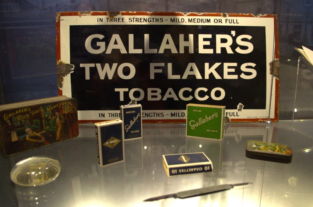 A display featuring vintage tobacco products. Central is a "Gallaher's Two Flakes" sign. Around are various cigarette boxes. The tone is nostalgic and historical.