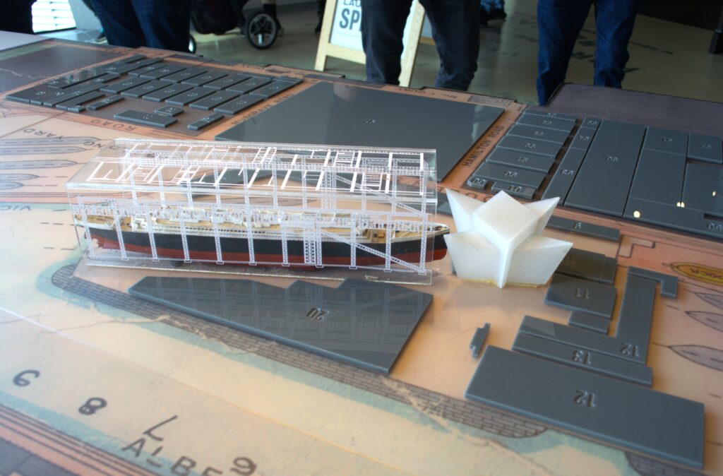 A detailed table display shows a transparent 3D-printed ship model with visible scaffolding. Gray panels with grid layouts are nearby. People stand in the background.