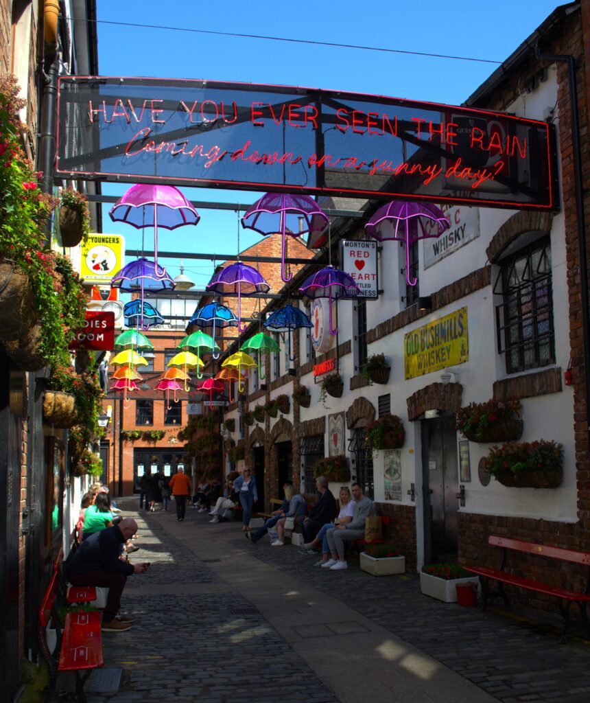 A narrow cobblestone alley adorned with colorful umbrella art overhead. Neon sign reads "Have you ever seen the rain?" People sitting on benches, vibrant atmosphere.
