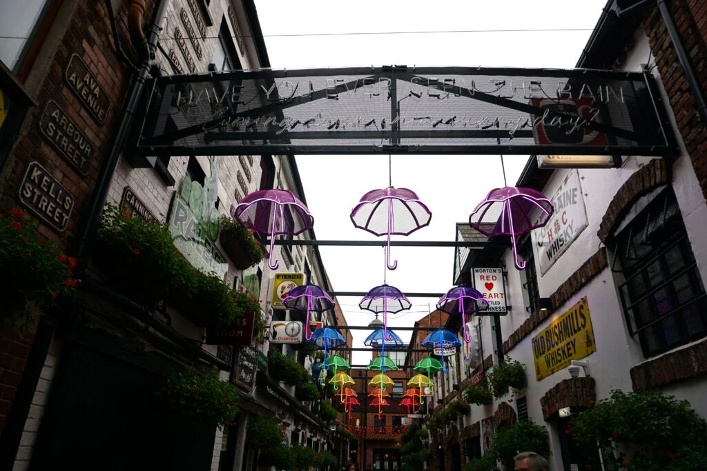 A narrow alley in Belfast is decorated with glowing neon umbrellas in purple, blue, green, yellow, and red hanging overhead. Vintage pub signs and flower boxes line the brick walls, creating a lively and colorful atmosphere. The vibrant lights and historic details capture the charm of Belfast’s pub lanes.