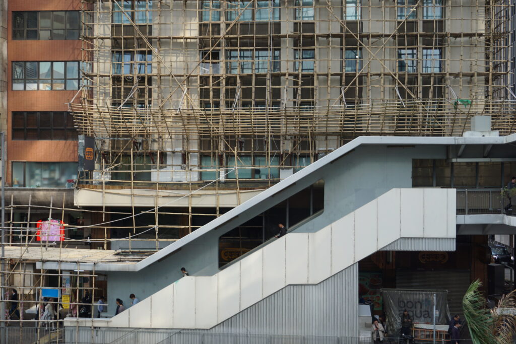 A busy urban scene in Hong Kong with pedestrians on a white pedestrian bridge, set against a tall building covered in bamboo scaffolding. The image conveys city life and construction.
