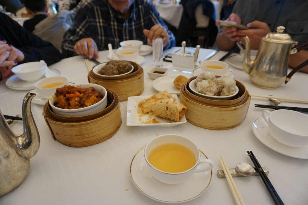A table with dim sum, featuring bamboo steamers of dumplings and buns, tea served in cups, and teapots. People are seated, creating a cozy atmosphere.