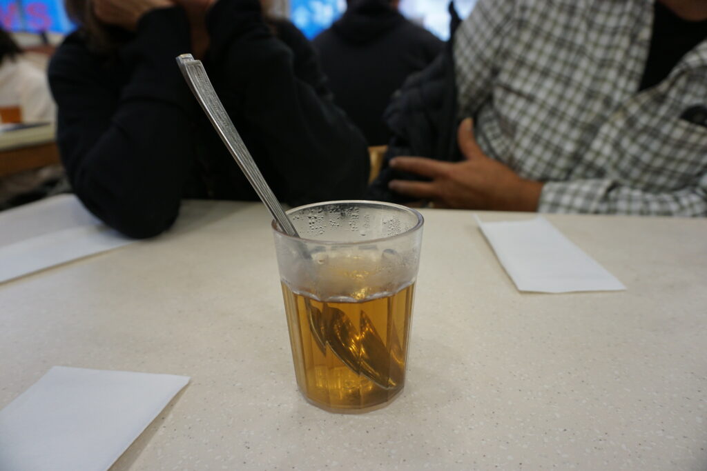 Clear plastic cup of hot tea with a metal spoon inside sitting on a light colored table in a casual restaurant. Two people sit blurred in the background making the warm amber tea the focal point of the image.