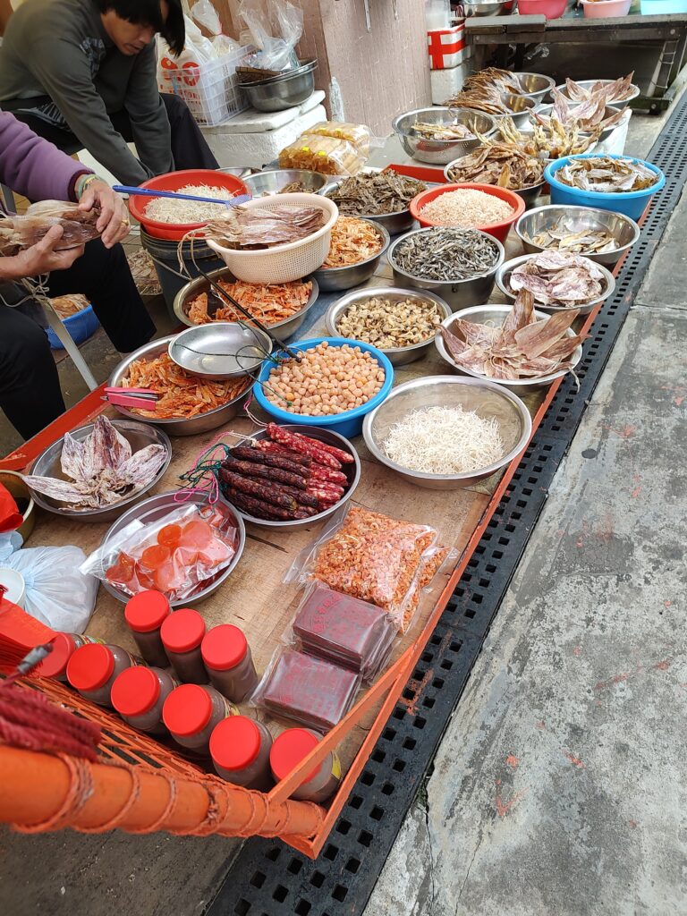 Street food market stall in Tai O featuring dried seafood, meats, and preserved goods in bowls and jars.