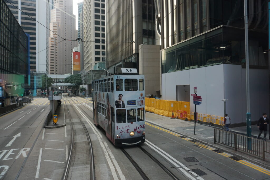 A Hong Kong ding ding, Tram number 86, travels along a street flanked by tall buildings in an urban setting. Yellow barricades and a few pedestrians are visible.