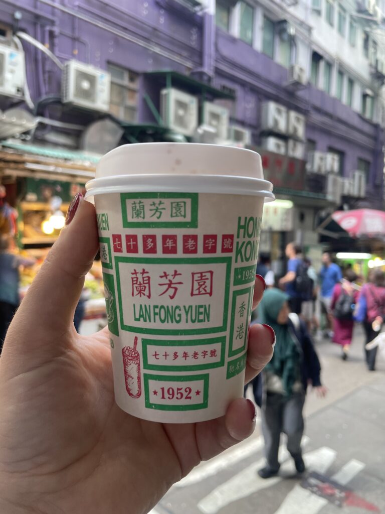 A hand holds a takeaway cup from Lan Fong Yuen in a bustling Hong Kong street. The purple building and busy market ambiance create a lively urban feel.
