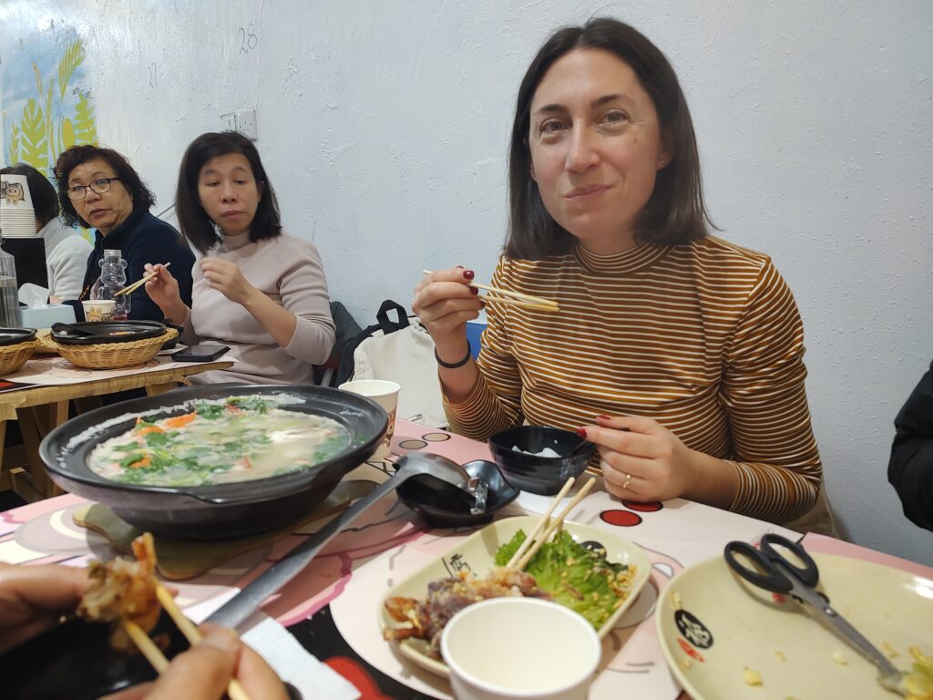 The picture depicts a restaurant scene. In the foreground, a woman with shoulder-length dark hair in a brown and white striped shirt is holding chopsticks and smiling slightly. In front of her are a small bowl and a dish with greens and chopsticks resting on it. To her left, another woman in a beige top is also holding chopsticks and looking slightly to her right. The table is cluttered with various dishes, including a large, black bowl filled with soup that contains vegetables and appears to be the central dish. Other items on the table include a pair of scissors, a plate with remnants of food, and paper cups. The background features a wall with a light blue and white textured surface and a partial mural of yellow and green plant motifs.