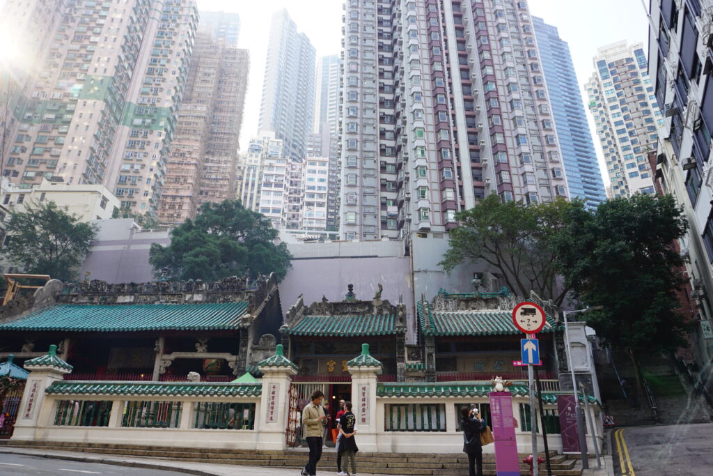 Traditional temple with green tiled roofs in foreground, set against towering modern skyscrapers. People walk nearby. The scene is busy yet serene.