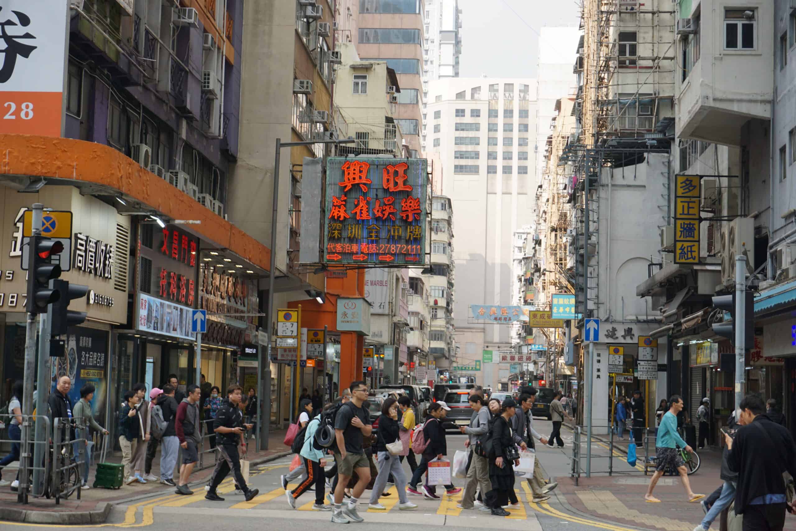 Bustling city street in Hong Kong, with busy pedestrian crossing, tall buildings with signs in Chinese, and a lively, vibrant urban atmosphere.
