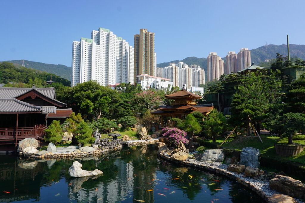 Serene Japanese garden with koi pond, lush greenery, and traditional wooden structures. Towering modern buildings rise in the distant background.