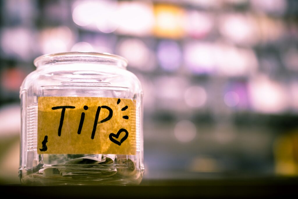 A clear glass jar labeled "Tip" in bold, black letters, decorated with a small heart. It contains cash, set against a blurred, colorful background.