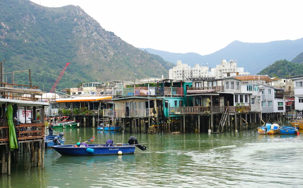 Stilt houses line the water in Tai O fishing village in Hong Kong with small boats floating in the canal. The homes stand on wooden poles with balconies and ladders against a backdrop of green mountains.