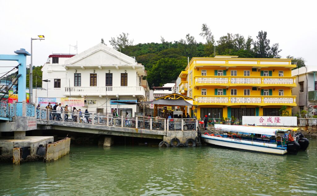 A waterfront view of Tai O fishing village in Hong Kong with colorful low rise buildings beside a canal. People gather on a pedestrian bridge while a small boat is docked in the water highlighting the village as an active fishing and tourist area.