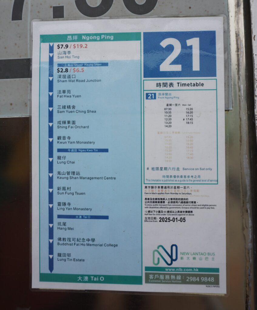 A bus stop sign for route 21 in Tai O Hong Kong shows destinations and fares in Chinese and English. The timetable lists service from Ngong Ping with departure times and notes it is operated by New Lantao Bus.