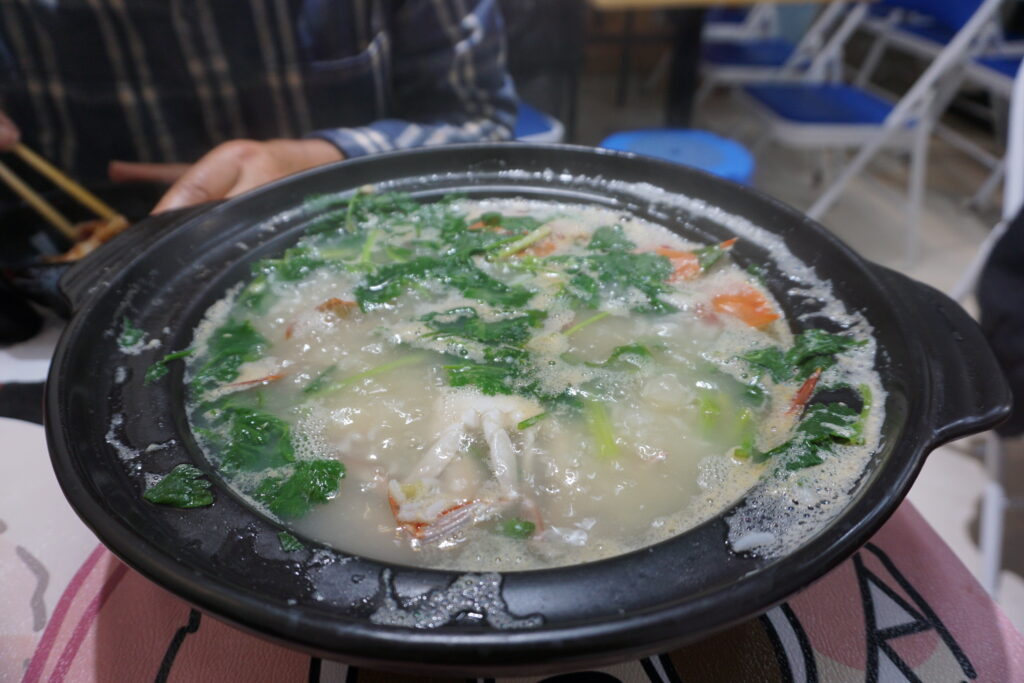The image depicts a close-up of a bubbling hot pot filled with various ingredients. The pot is black and circular, with condensation and frothy bubbles visible around the edges, indicating heat. Inside the pot, there are leafy greens, reddish-orange crab pieces, and other indistinct ingredients submerged in a cloudy, broth-like liquid. In the background, there's a faint view of a person holding chopsticks, wearing a plaid shirt, and a setting with blue folding chairs and tables, suggesting a casual dining environment.